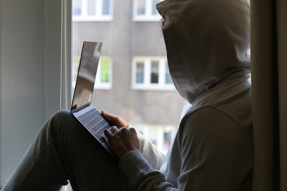 A person wearing a hoodie sits on a window sill, working on a laptop